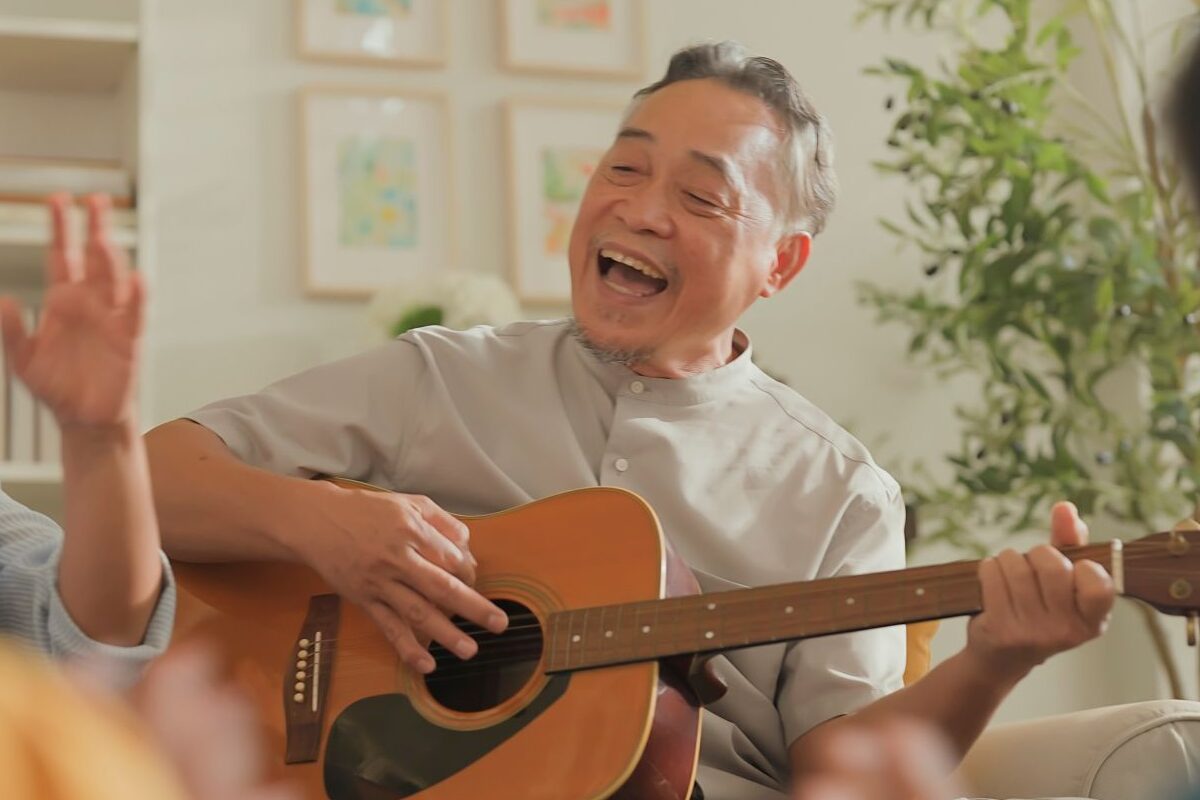 An older Asian man joyfully plays an acoustic guitar and sings in a cozy indoor gathering, showcasing the vibrant community life of Independent Living in Boise.