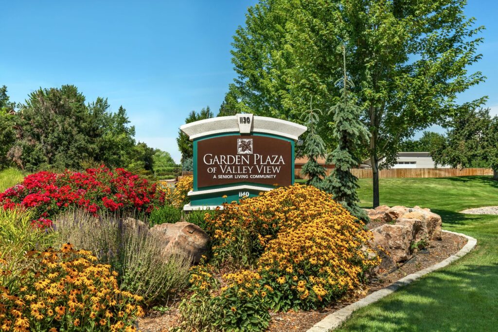 Senior living in Boise highlighted by the landscaped entrance sign for Garden Plaza of Valley View, surrounded by mature trees, colorful flowers, and a well-maintained green lawn.