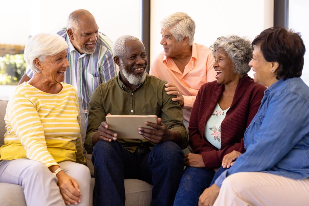 A diverse group of seniors laughs and gathers together around a tablet, showcasing the joy and connection that come with the benefits of senior friendships.