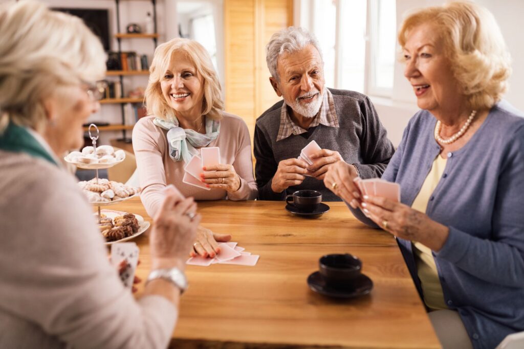 A group of older adults laugh and play cards together around a wooden table, capturing the connection and cognitive engagement at the heart of the social benefits of games for seniors.